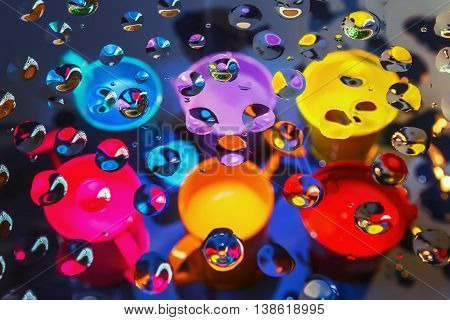 Bright abstract background - view through glass with drops of liquid on a blurry colorful watering cans and buckets. Selective focus on foreground.