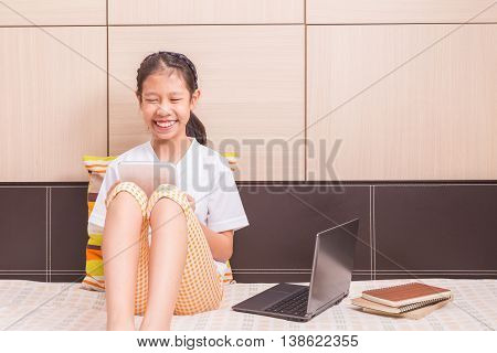 Happy smiling asian girl using tablet computer to study on her bed with laptop notebook computer and paper notebooks sitting on her bed room for copy space text