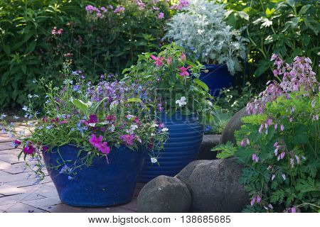 Bright blue pots overflow with colorful flowers in a shaded brick patio.