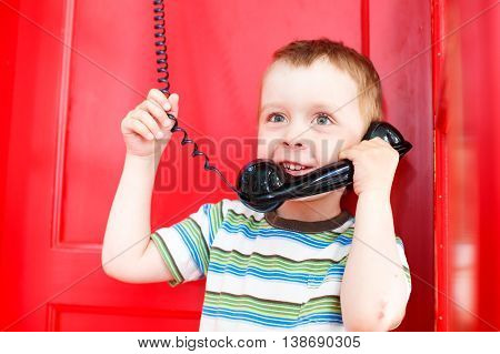 child holding a telephone receiver and smiling. cute boy talking on the phone in a red telephone box. the concept to share with the family joy and impressions