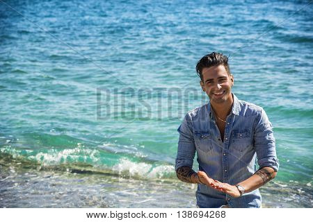 Handsome Athletic Young Man in Trendy Attire, on a Beach in a Sunny Summer Day, Looking At Camera, against Blue Sea Background.