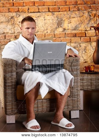 ¿Retrato de hombre con albornoz usando la computadora portátil en el sillón, relajarse en el bienestar.?