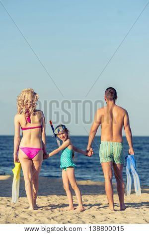 Back view of young parents and their little daughter in swimsuits holding hands standing on the sunny beach. Couple is holding swim fins. Girl is looking at camera