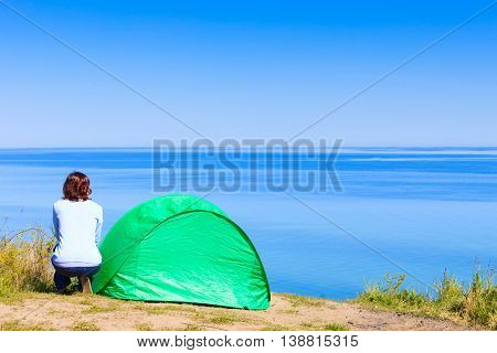 Tourism and recreation. Small green tent in natural area surrounded by meadow water sea lake. Woman spending time on fresh air.