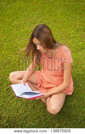 Beautiful young girl reading a book sitting on the green grass