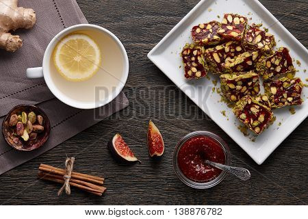 White cup of ginger tea with lemon, pistachio nuts, fig jam and turkish delight on a dark wooden background