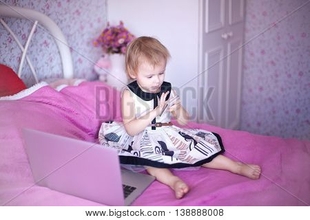 baby in pink room sitting on the bed and playing or watching cartoons price. Girl sitting at home. little princess in dress uses advanced fashion technology