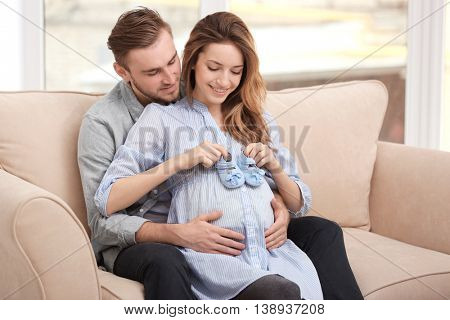 Happy pregnant woman holding baby shoes  and her husband on a sofa at home