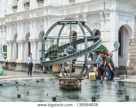 MACAU CHINA - MAY 24: Tourists and local residents walk pass the metal glope statue in the fountain at the Senado Square on May 24 2016 in Macau China.