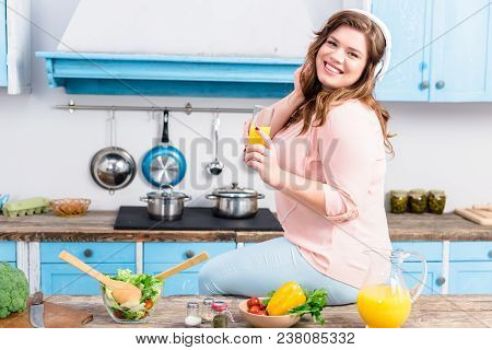 Smiling Overweight Woman In Headphones With Glass Of Juice In Hand In Kitchen