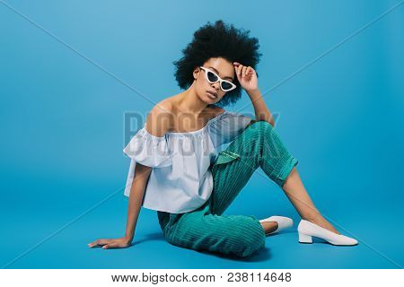 Attractive Young Woman In Stylish Off-the-shoulder Top And Sunglasses Sitting On Floor On Blue