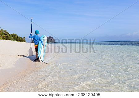 Back View Of Little Boy In Rashguard Holding Surf Board And Paddle Walking At The Beautiful Fiji Isl