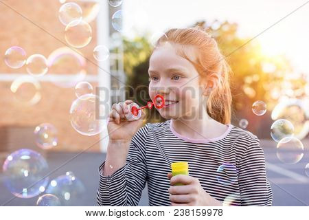 Portrait of lovely little girl with red hair blowing soap bubbles at park. Portrait of cute girl blowing soap bubbles outside the school. Happy female child playing with soap bubble at playground.
