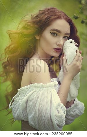 A Sensual Red-haired Girl In A White Dress With Bare Shoulders Holds A White Dove In Her Hand. White