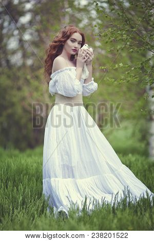 A Sensual Red-haired Girl In A White Dress With Bare Shoulders Holds A White Dove In Her Hand. White