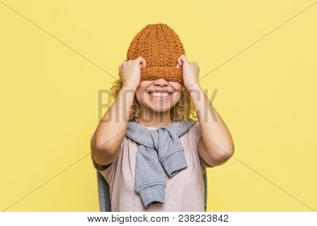 Happy Girl Is Stretching Hat Down. She Has Put It Onto Her Yeys And Smiling. Isolated On Yellow Back