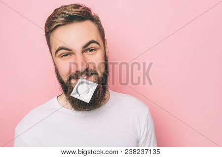Happy Man Is Looking Straight Forward And Holding A Condom With His Lips. Isolated On Pink Backgroun