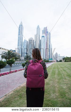 Beautiful Girl With A Backpack Looking At The Tall Beautiful City Buildings Near The Road
