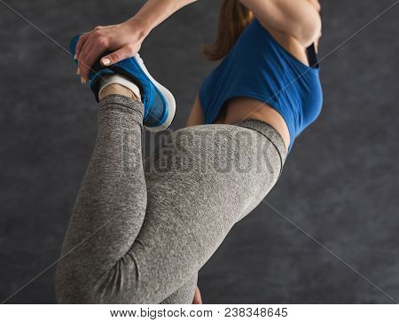 Young Woman Warming Up Before Training At Gym. Unrecognizable Girl Stretching Her Leg At Gray Backgr