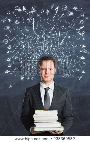 Businessman Holding Books On Abstract Chalkboard Background With Drawin Arrows. Choice And Education
