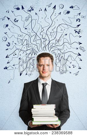 Businessman Holding Books On Abstract Concrete Background With Drawin Arrows. Choice And Education C