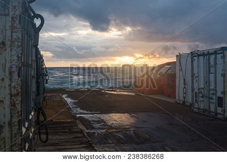 View On Cargo Deck With Containers Of Offshore Supply Vessel. Sunset, Oil Rigs Are On Background