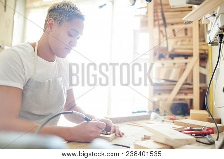 Young carpenter making decorative details from wood while carving with special pyrographic machine