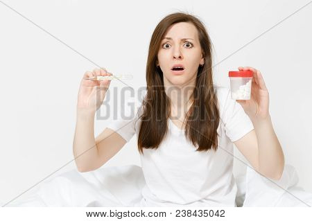 Young Sick Tired Sad Woman In Pajamas Sitting On Bed, Holding Clinical Thermometer With High Fever T