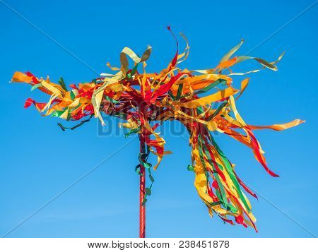 Festive Wooden Pole For Climb On Maslenitsa. Wooden Wheel With Colorful Ribbons On Blue Sky Backgrou