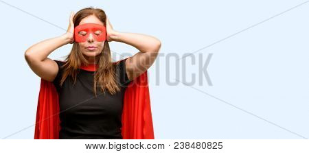 Middle age super hero woman wearing red mask and cape covering ears ignoring annoying loud noise, plugs ears to avoid hearing sound. Noisy music is a problem. isolated blue background