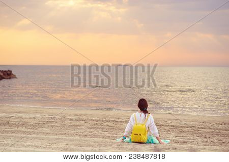 Young Woman With Yellow Backpack Sitting On Sandy Beach Looking At Sea And Sunset. Adriatic Sea In D
