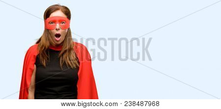 Middle age super hero woman wearing red mask and cape scared in shock, expressing panic and fear isolated blue background