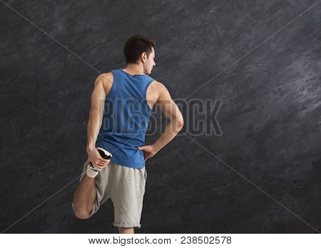 Young Man Warming Up Before Training At Gym. Guy Stretching His Leg At Gray Background Indoors, Copy