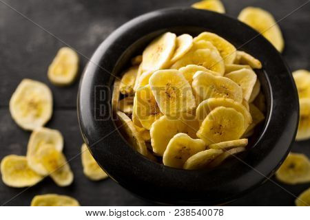Heap Of Dried Banana Chips Snack In Black Bowl On Dark Rustic Table With Selective Focus