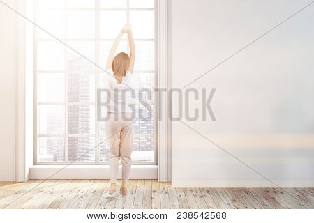 Woman In Pajamas In An Empty Room With White Walls, A Wooden Floor, A Loft Window With A Cityscape A
