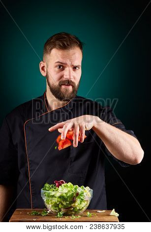 Portrait Of A Handsome Mischevious Male Chef Cook In Black Uniform Making Delicious Vegetable Salad 