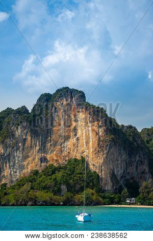 A White Sail Boat On A Calm Emerald Water In Front Of A Limestone Mountain At Railay Beach At Krabi,