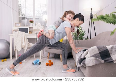 In High Spirits. Determined Athletic Mother Training At Home And Holding Her Daughter On Her Back