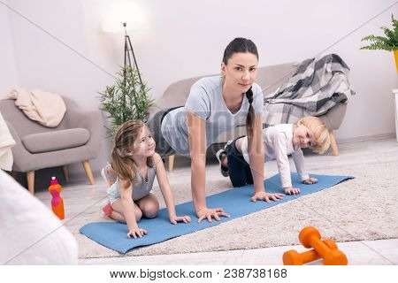 Home Trainer. Content Young Mother Smiling And Doing Push-ups With Her Kids