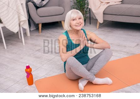 Mood-boosting Exercise. Upbeat Elderly Woman Sitting Cross-legged On A Yoga Mat And Smiling At The C