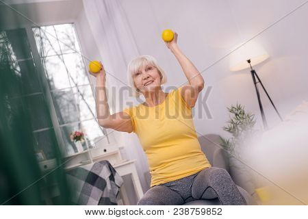 Upbeat Mood. Beautiful Elderly Woman Smiling Pleasantly And Lifting Up Dumbbells While Working Out S