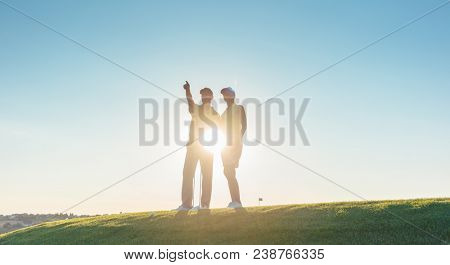 Low-angle view of the silhouette of a man pointing to the horizon, while standing next to his female partner on a professional golf course against sunshine and clear blue sky