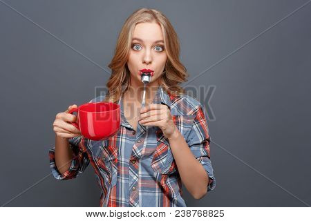 Amazed Girl Is Standing And Posing. She Is Holding A Red Cup In Her Hand And Keeping The Small Spoon