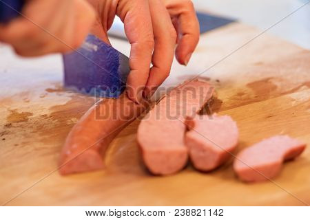 Hands Slicing Sausage Wih Blue Knife On Wood Cutting Board.