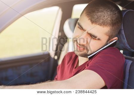 Close Up Portrait Of Focused Unshaven Male Driver Talks Via Cell Phone While Drives Car, Being In Hu