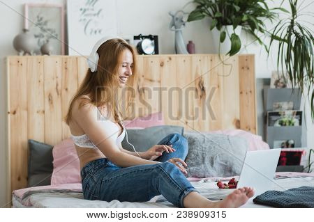 Young Sexy Blonde Woman Flirting Online On Laptop, Sitting On Bed In Bedroom. She Wearing Bra, Jeans