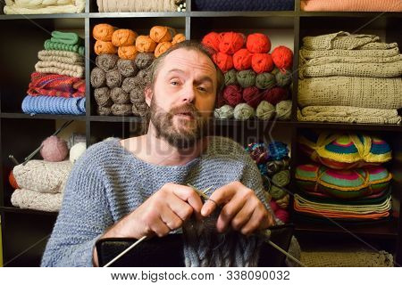 European Man 40-50 Years Old Knits A Scarf. A Man In A Knitted Sweater On The Background Of Shelves 