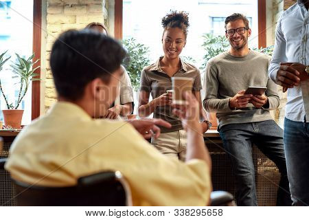 Portrait Of Young Male Office Worker In A Wheelchair Drinking Coffee And Chatting With Colleagues Du