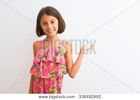 Young beautiful child girl wearing pink floral dress standing over isolated white background smiling cheerful presenting and pointing with palm of hand looking at the camera.