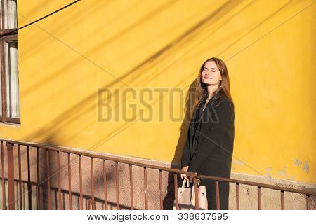 Beautiful Girl In Black Cloack And Handbag In Hands Leaned On The Yellow Wall And Bask In The Sun Wi